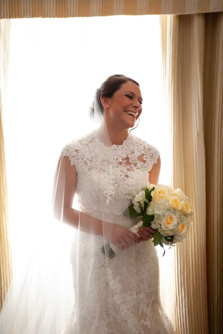 Joyful bride laughs while holding yellow rose bouquet in sunlit room.