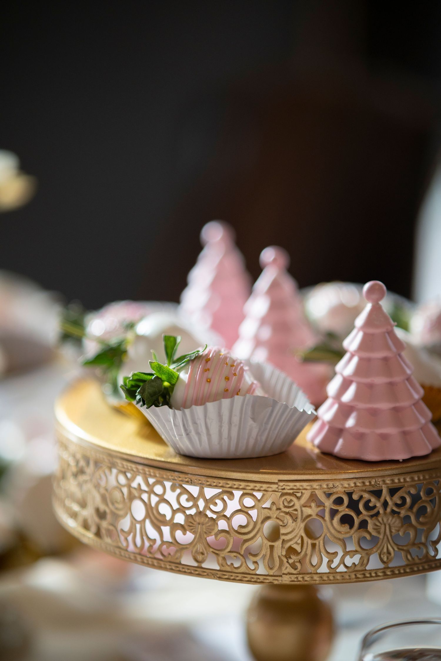 Pink holiday desserts and decorative trees displayed on an ornate gold cake stand with filigree details.