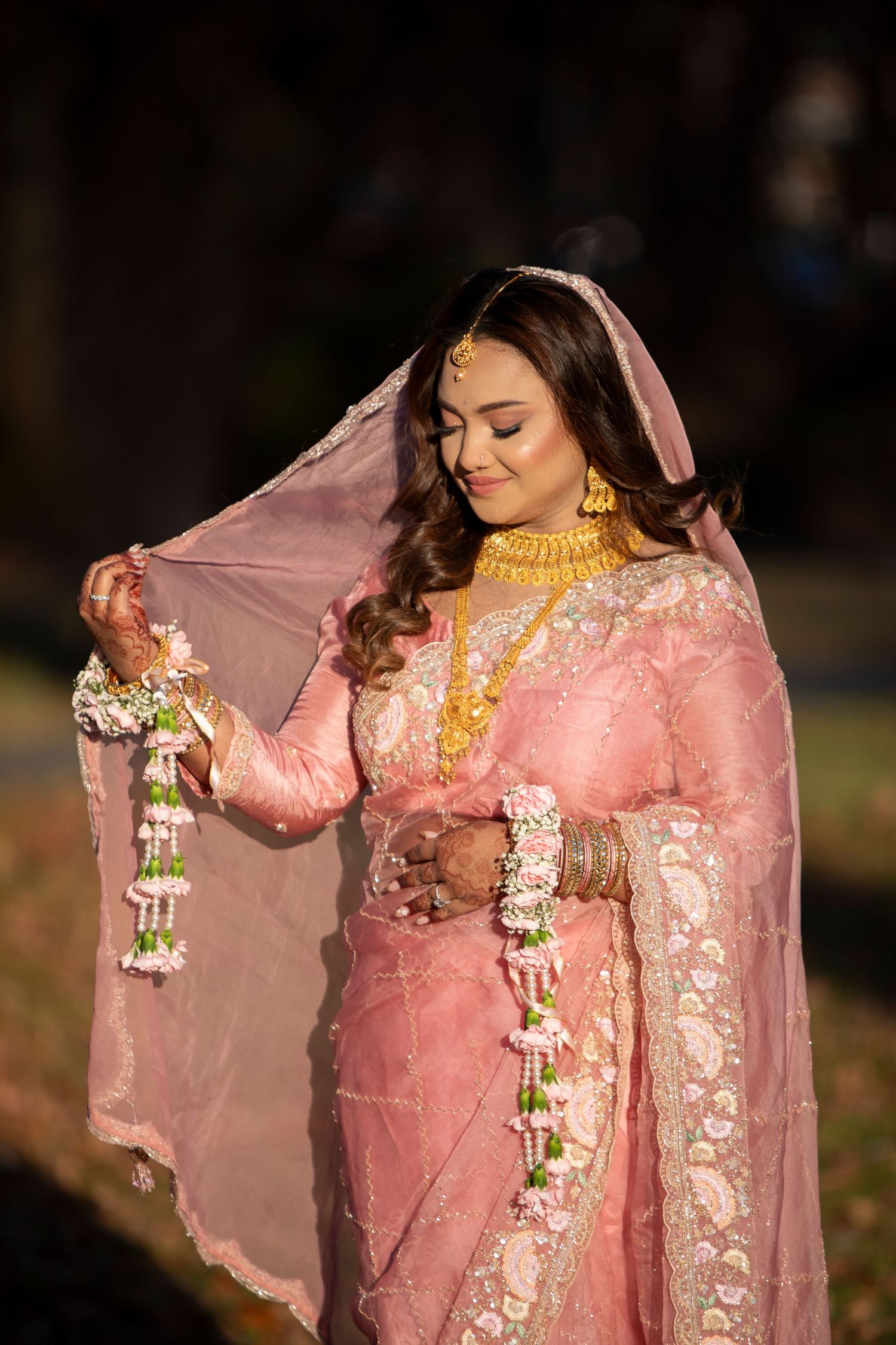 A bride in a pink and gold traditional South Asian wedding outfit adjusts her dupatta in outdoor lighting.