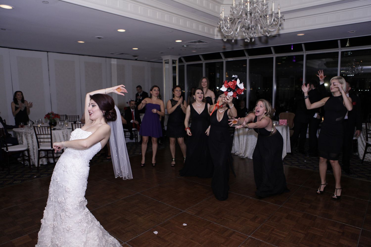 Bride in white mermaid dress tosses bouquet to waiting guests under crystal chandelier.