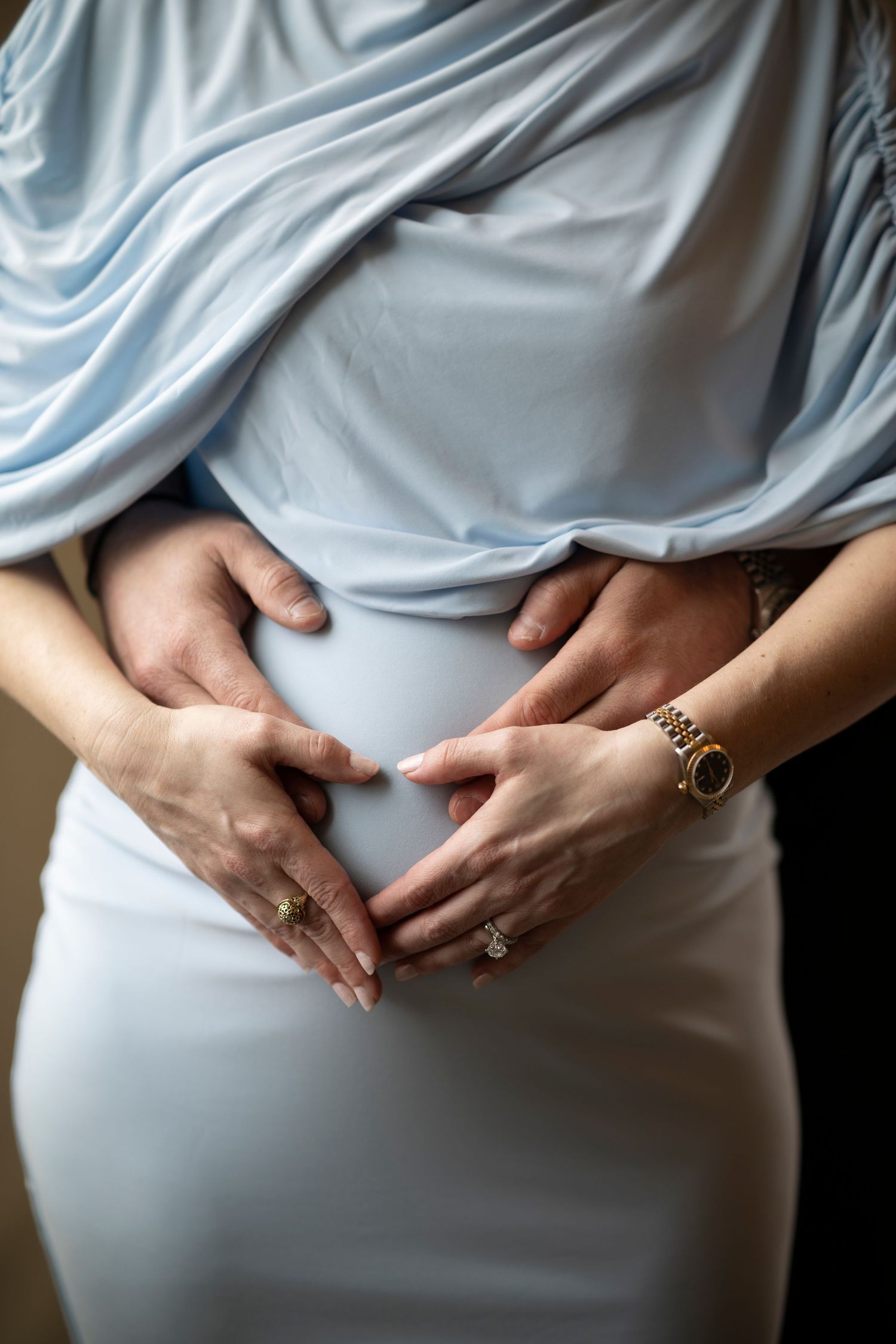 Hands form a heart shape over a pregnant belly draped in soft blue fabric with an engagement ring visible.