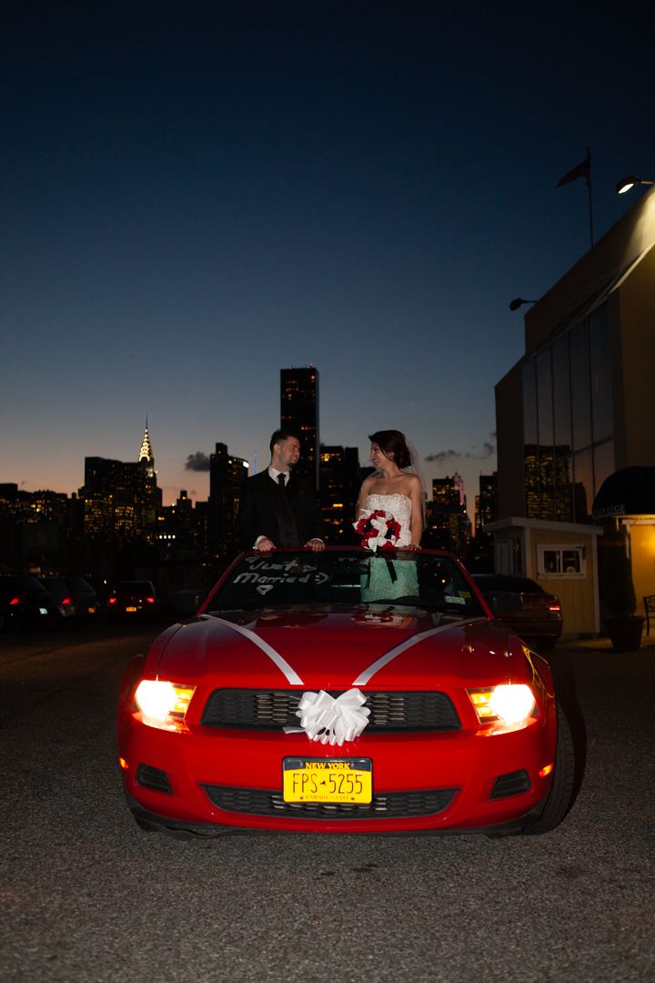 Couple poses through sunroof of red Mustang with decorative bow against nighttime city backdrop.