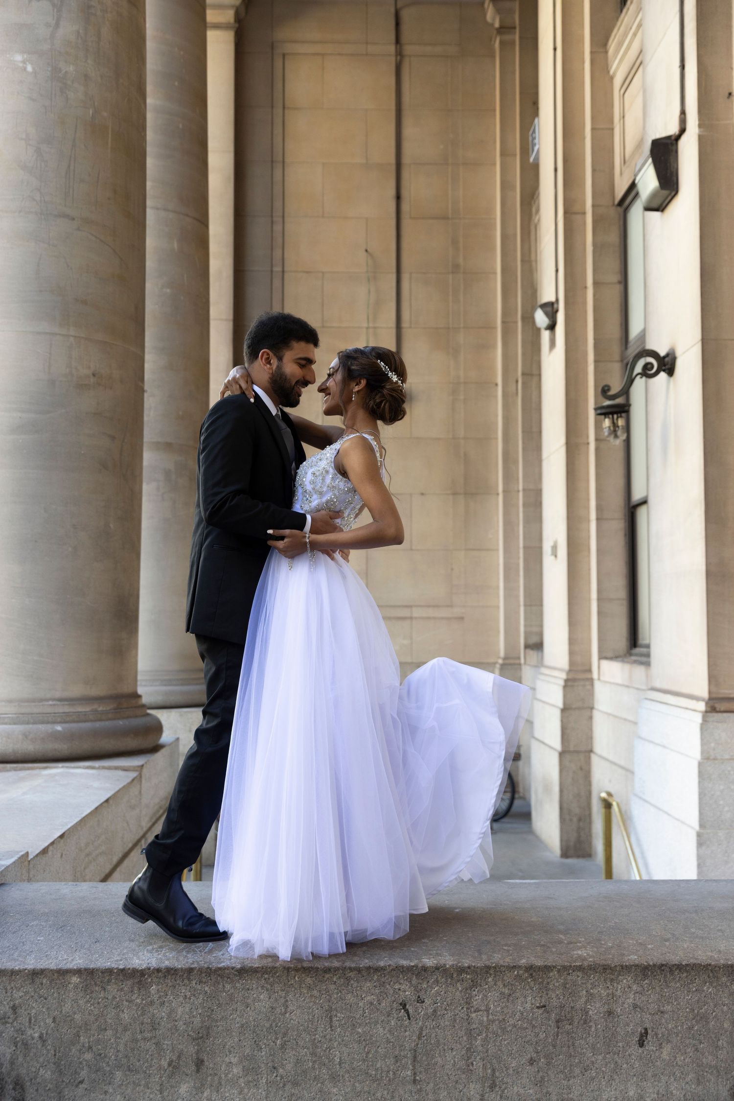 A romantic wedding couple shares a kiss in an elegant colonnade hallway with white marble columns.