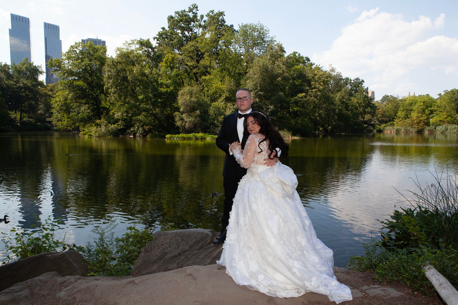 Wedding couple photographed against scenic Central Park lake with skyline in background.
