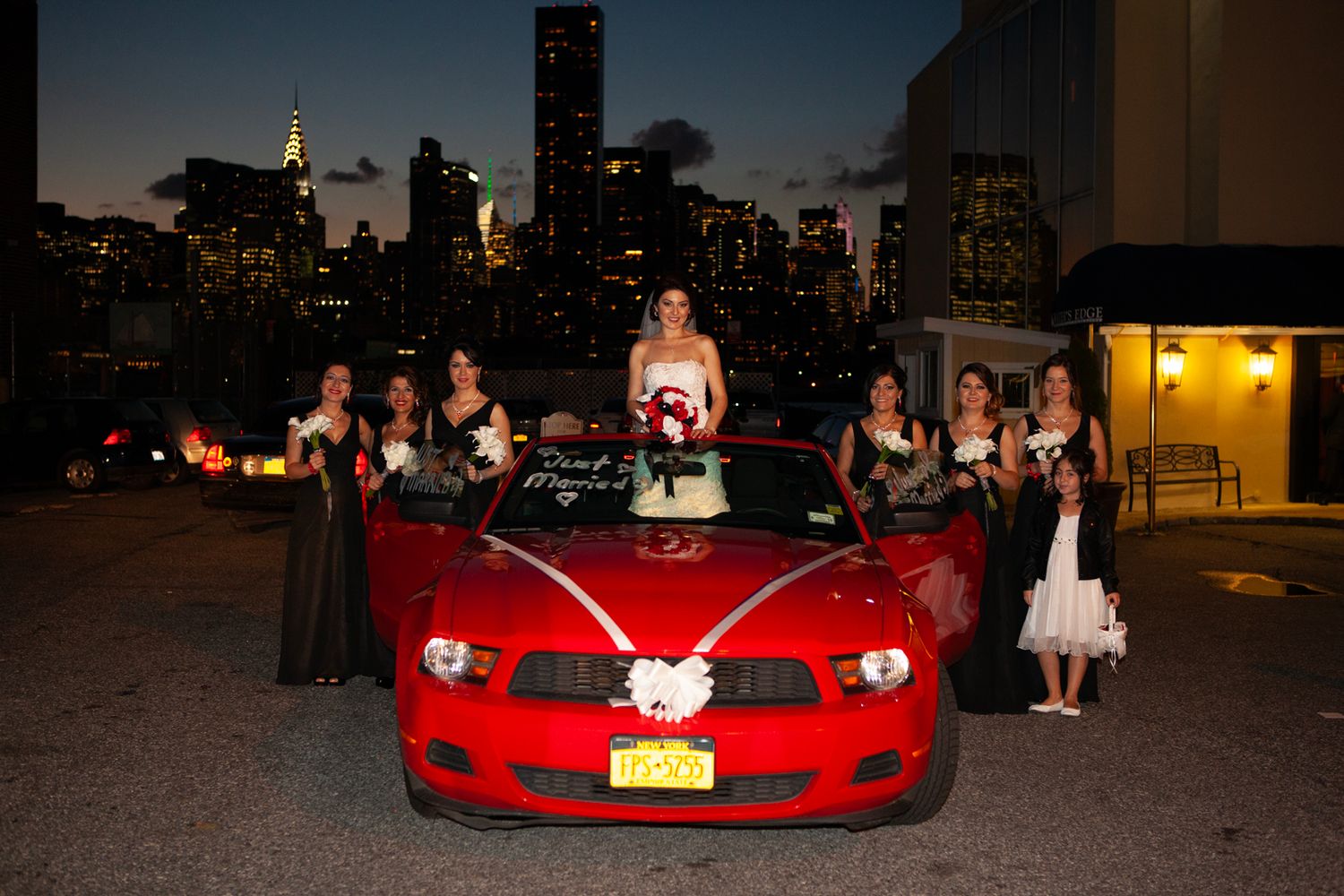 Wedding party poses with red sports car against New York City skyline at dusk.