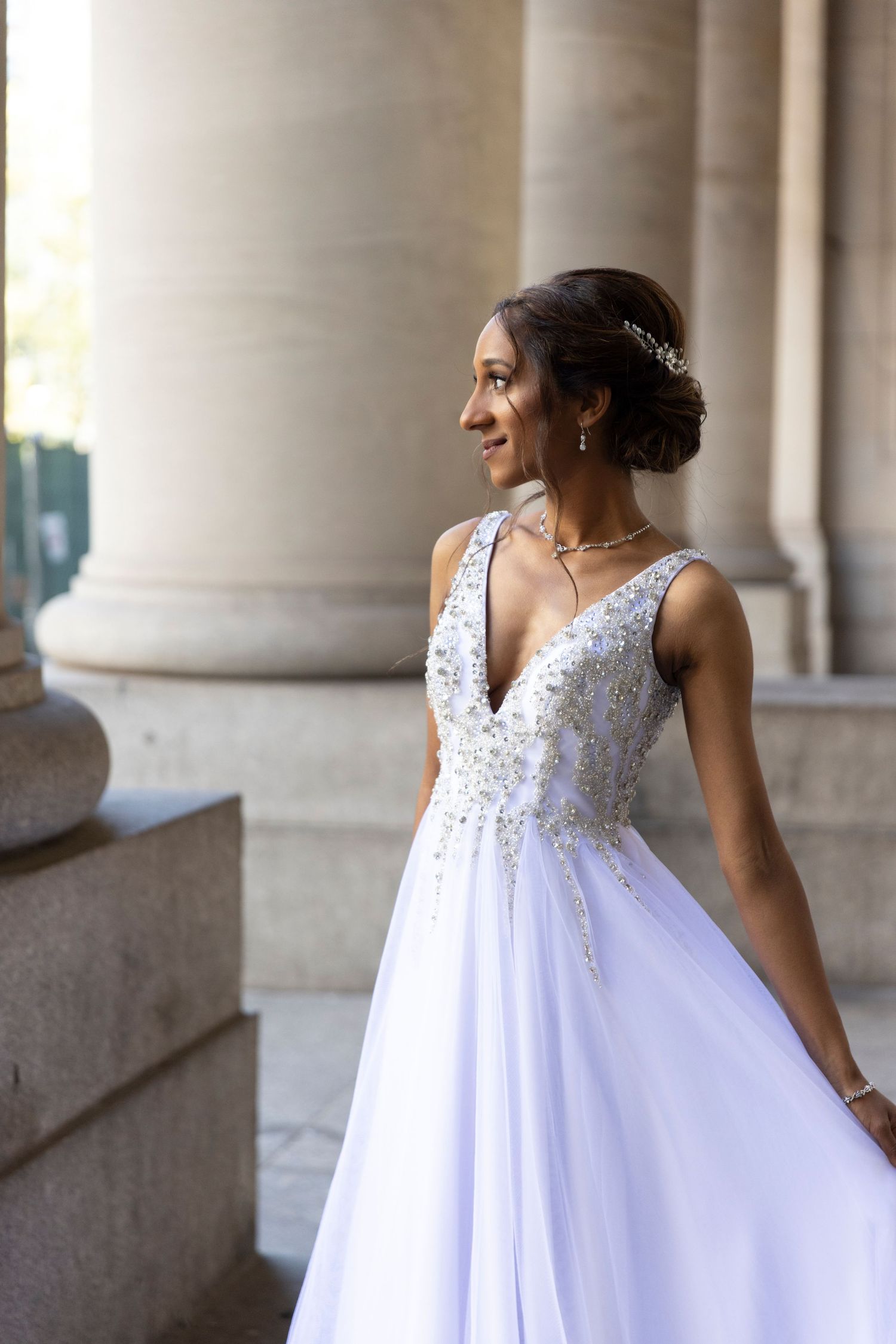 A bride in a flowing white wedding dress poses elegantly between classical stone columns in a romantic setting.