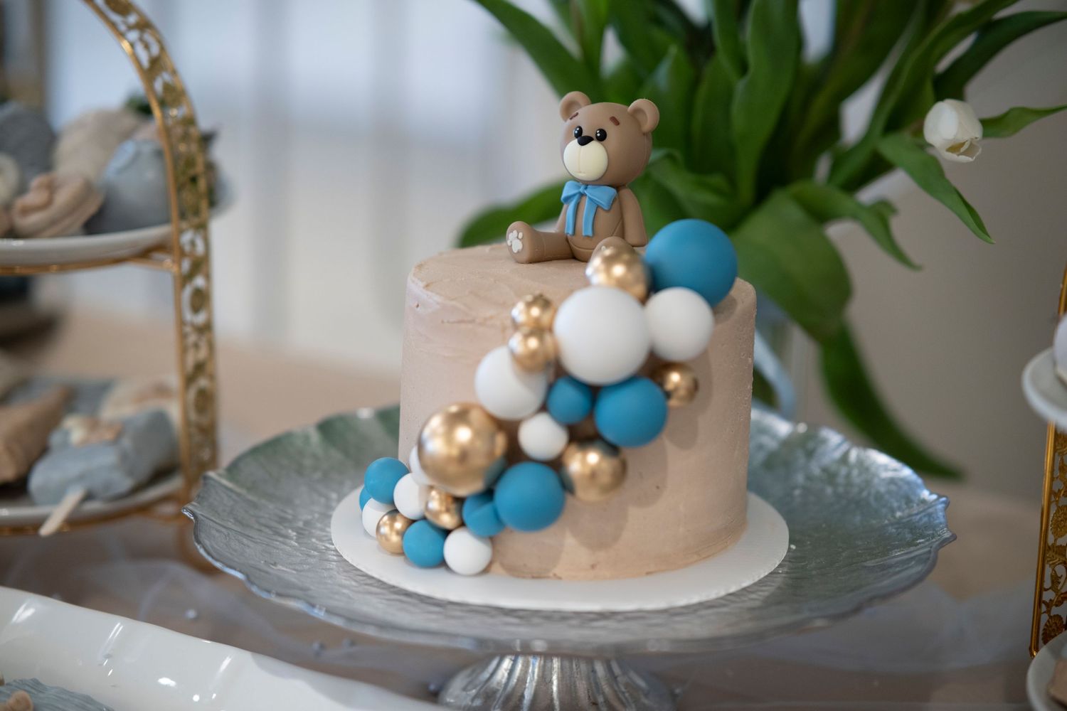 Elegant wedding cake decorated with a teddy bear topper and blue balloon accents on a crystal cake stand.