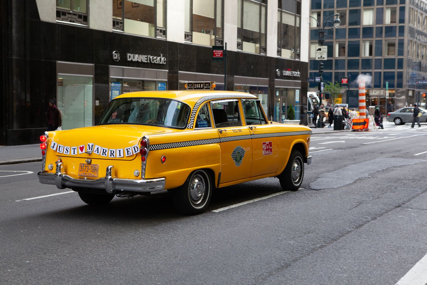 Vintage yellow taxi cab drives through a city street with buildings in the background.