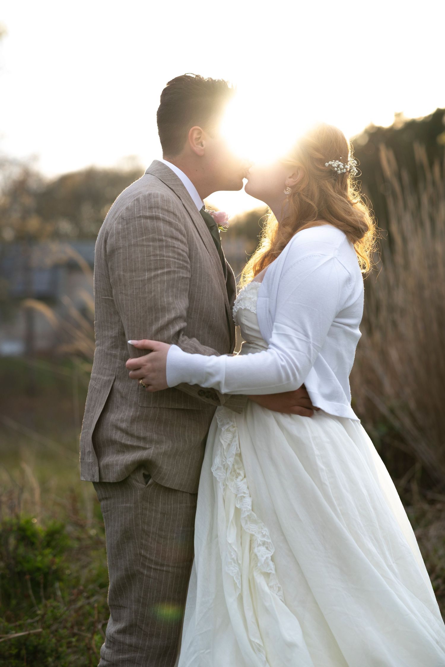 Bride and groom share a romantic sunset kiss outdoors, she in white lace gown, he in tan plaid suit.