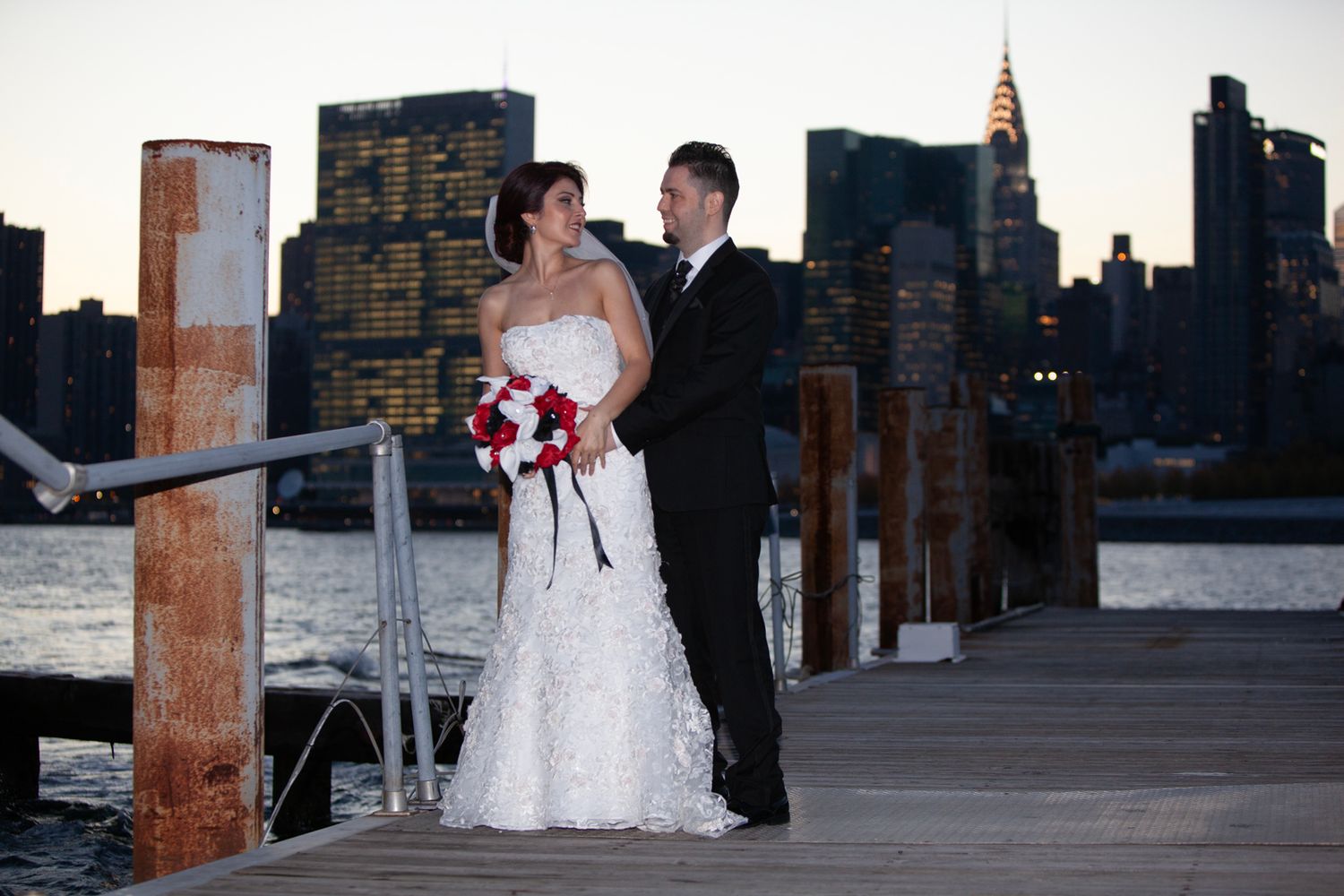 Newlyweds pose on wooden pier with NYC skyline backdrop at dusk.