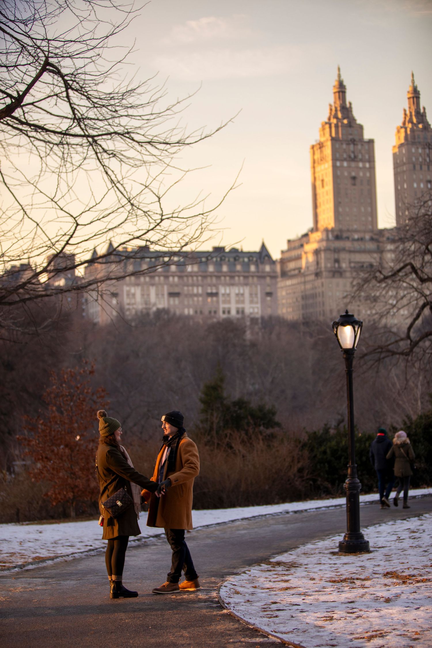 Two people enjoying a winter afternoon on a snowy path in Central Park with iconic San Remo apartment towers in background.