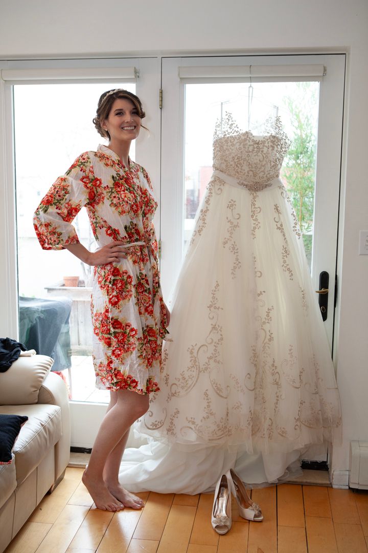 Woman in floral dress poses playfully next to elegant white wedding gown in bright room.