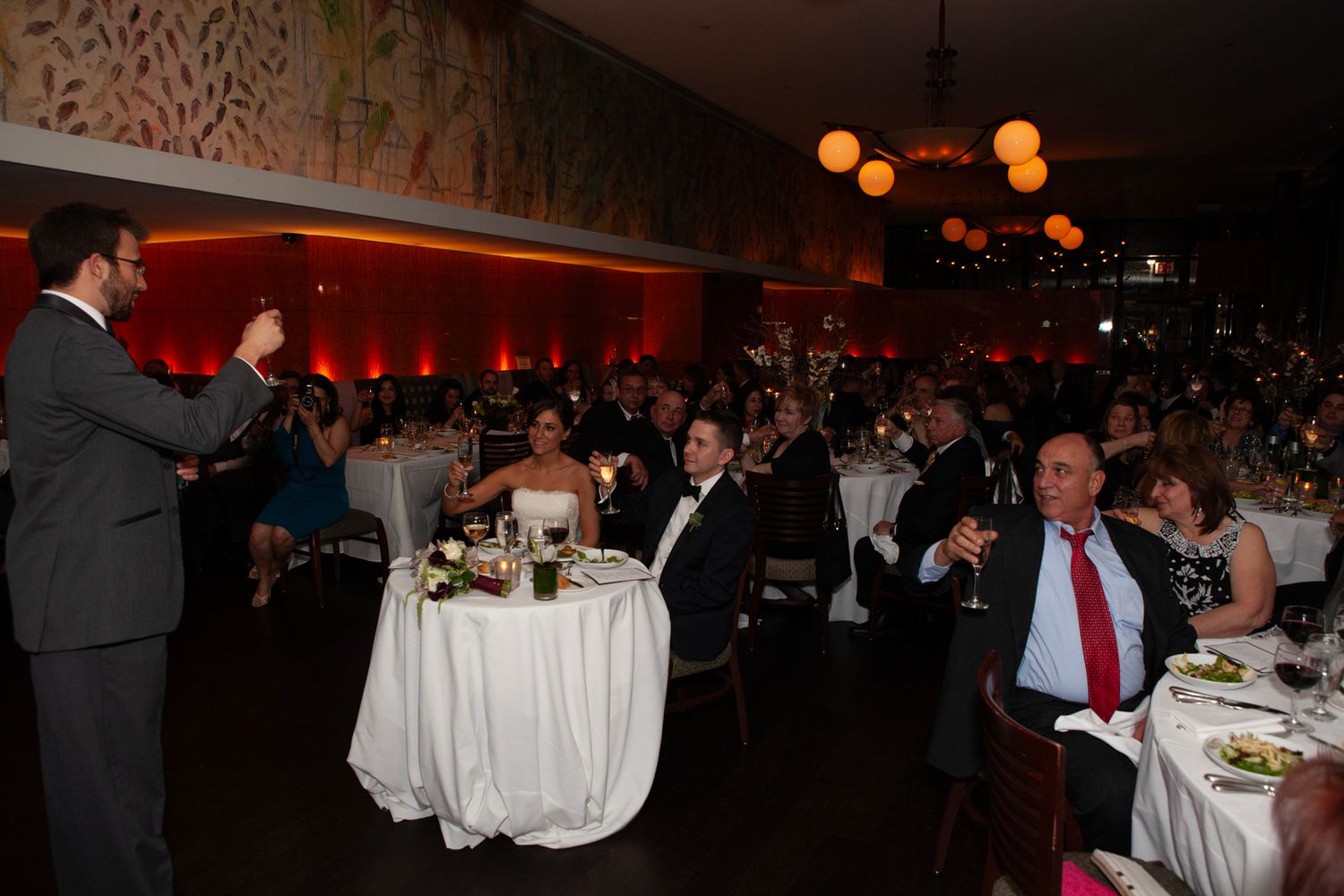 Wedding reception venue with guests seated at decorated tables and speaker addressing crowd.