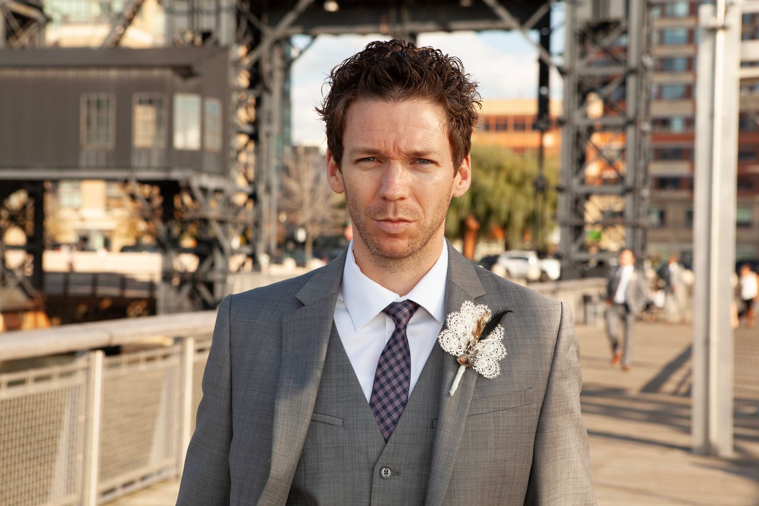 Groom in grey suit with boutonniere stands outdoors on sunny day.
