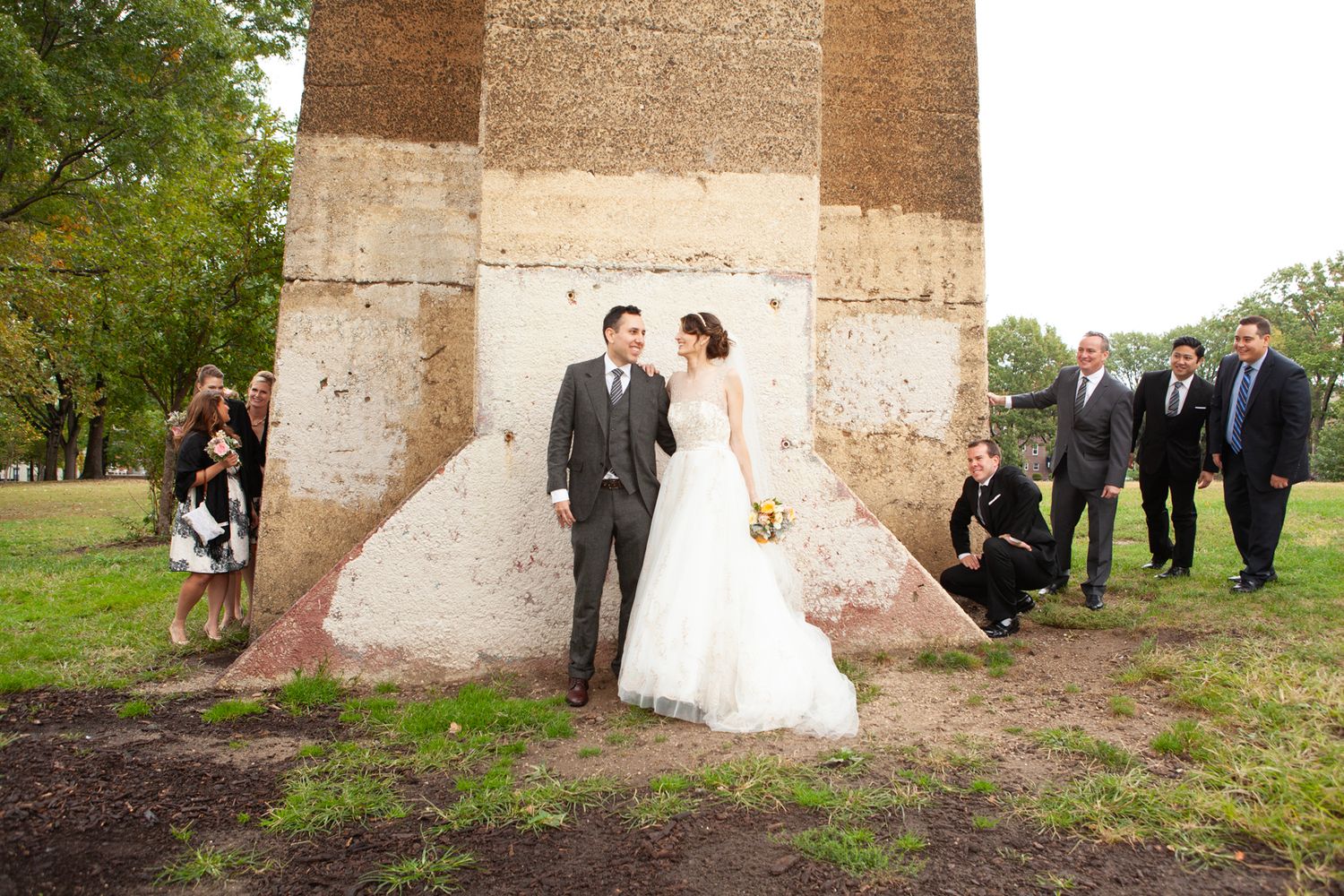 Wedding party poses against large stone bridge support on grass.