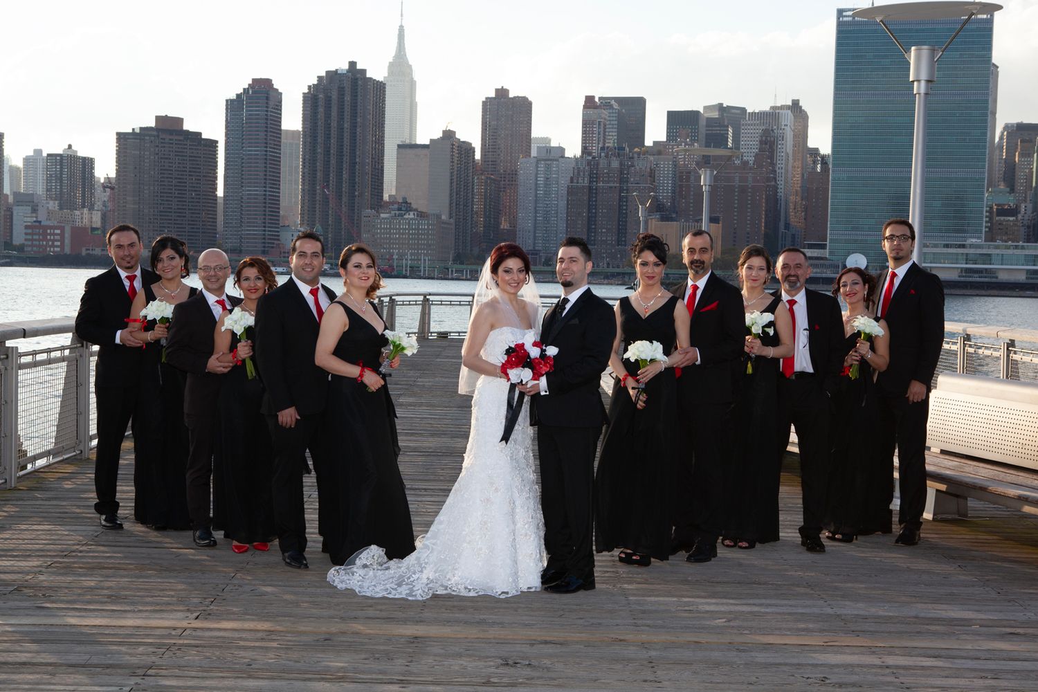 Wedding party in black formal wear poses on waterfront with city skyline backdrop.