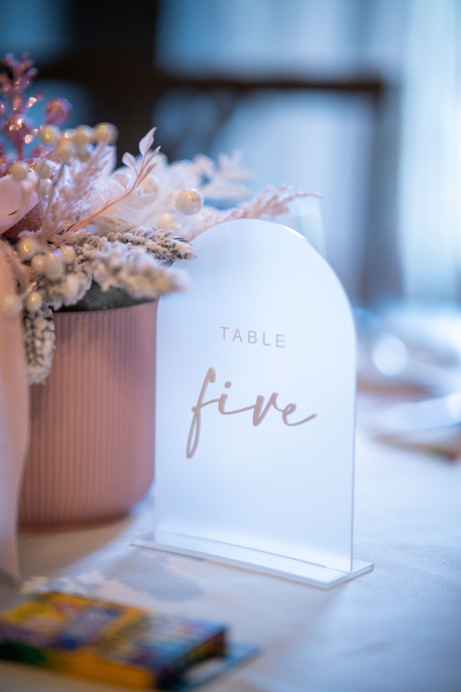 Elegant wedding table decor featuring pink floral centerpieces and white table number cards with gold lettering.