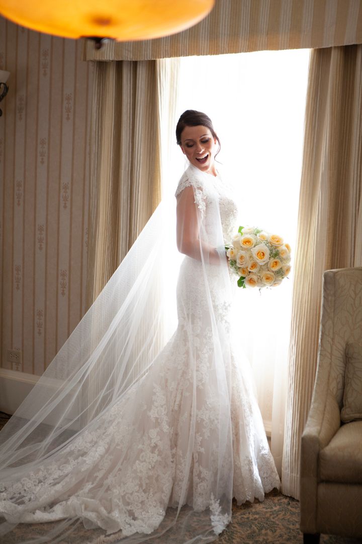 Radiant bride in lace wedding gown holds yellow rose bouquet by window.