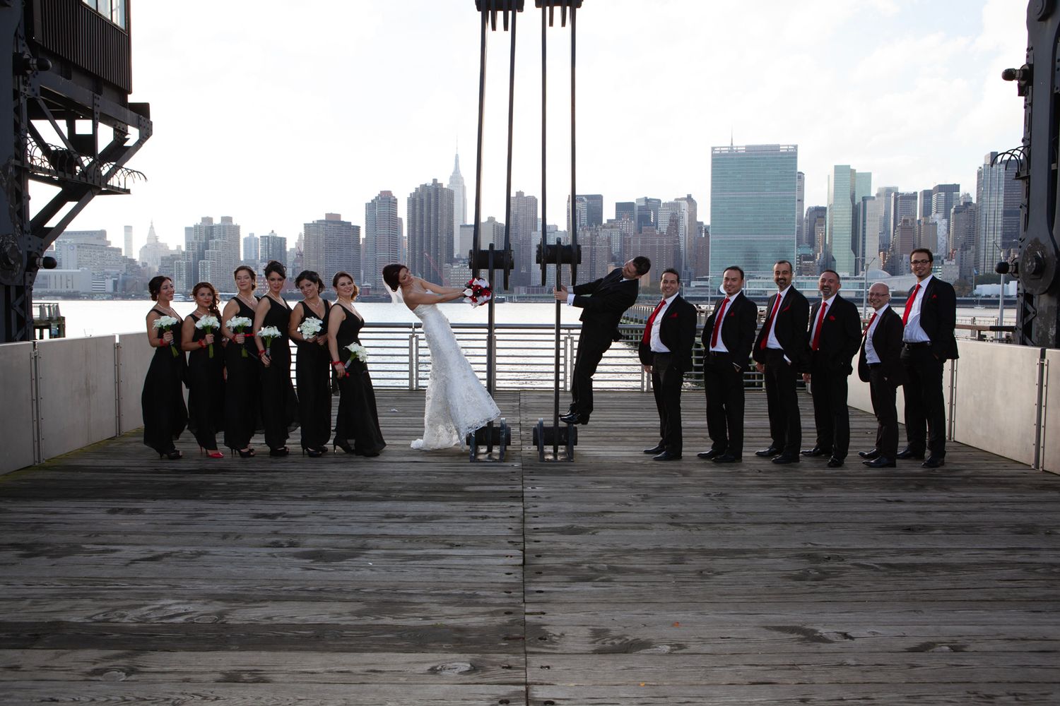 Wedding party split into two groups poses dramatically on a pier with city skyline backdrop.