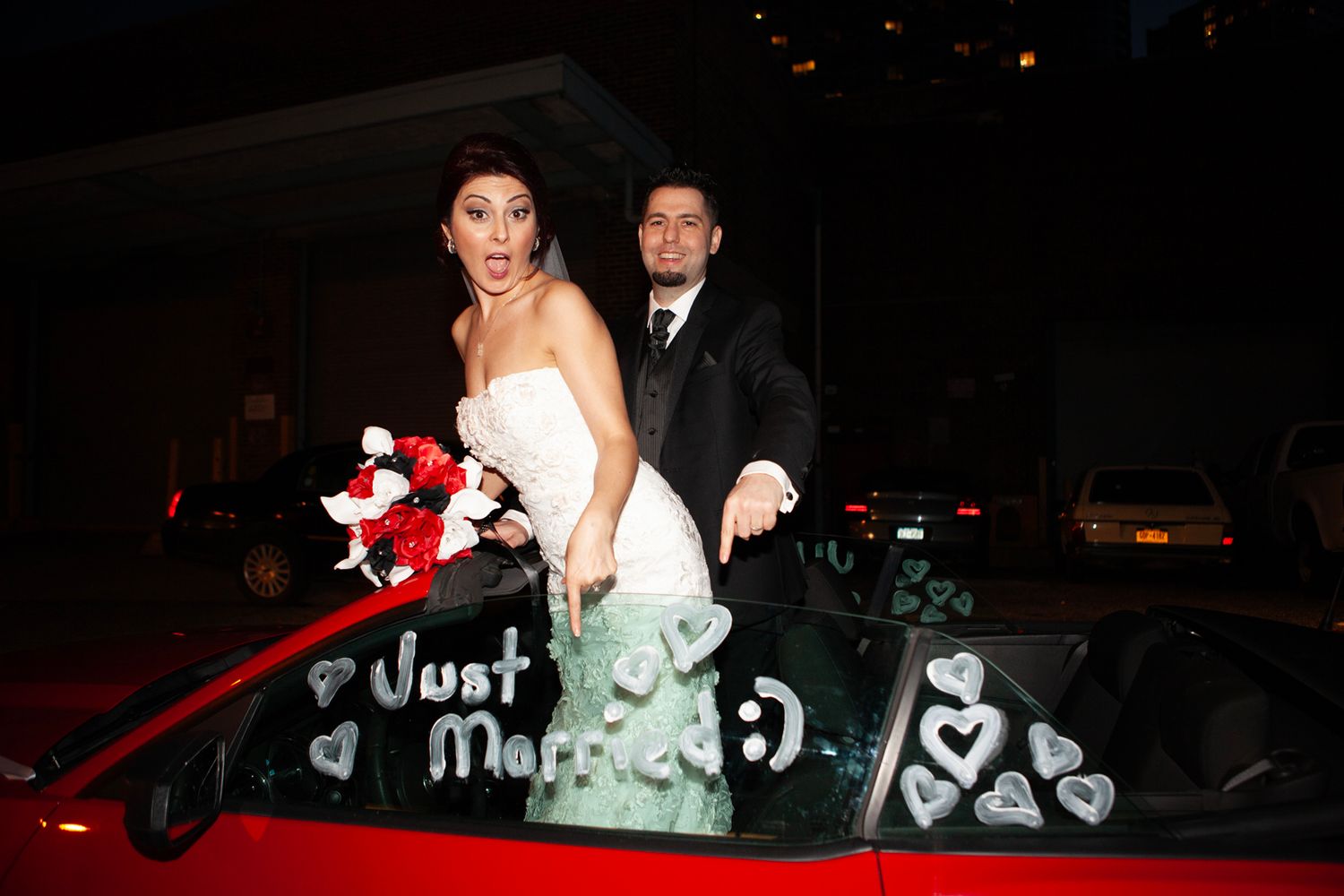 Newlyweds pose with red and white bouquet on decorated red car with 'Just Married' written on window.