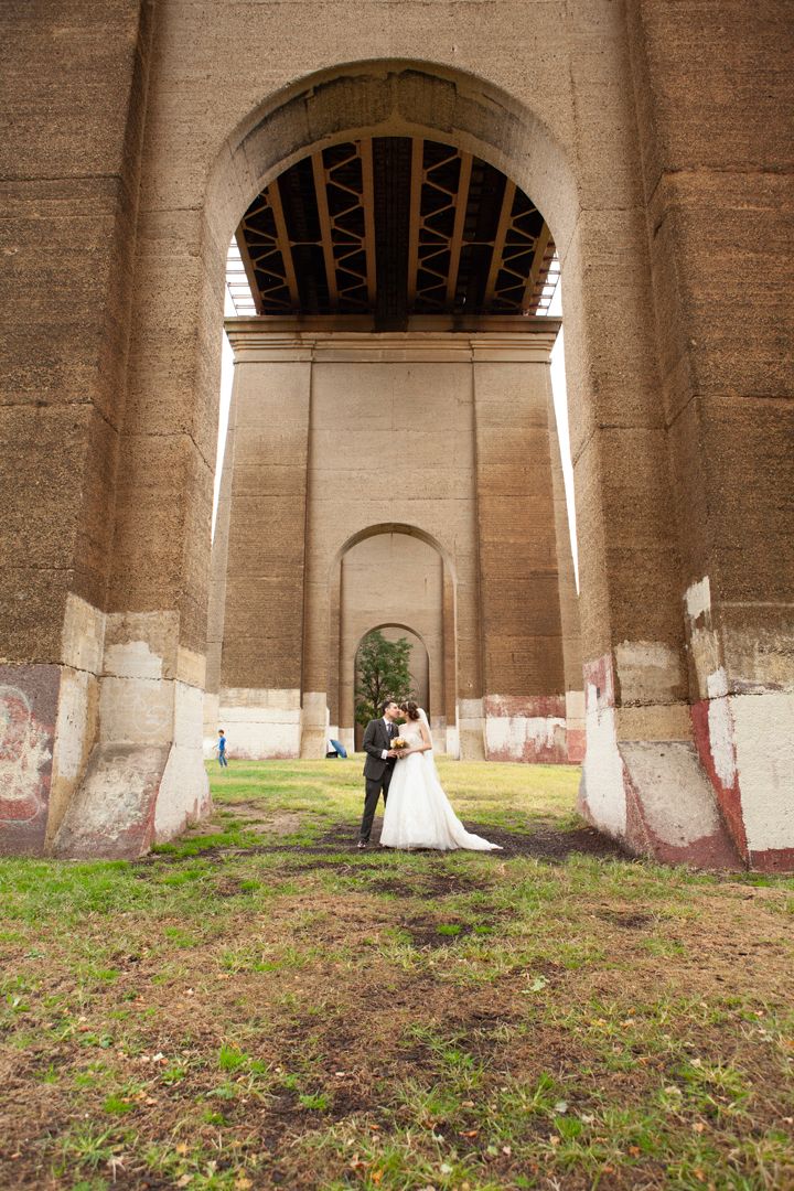 Wedding couple photographed beneath massive stone bridge supports on grassy ground.