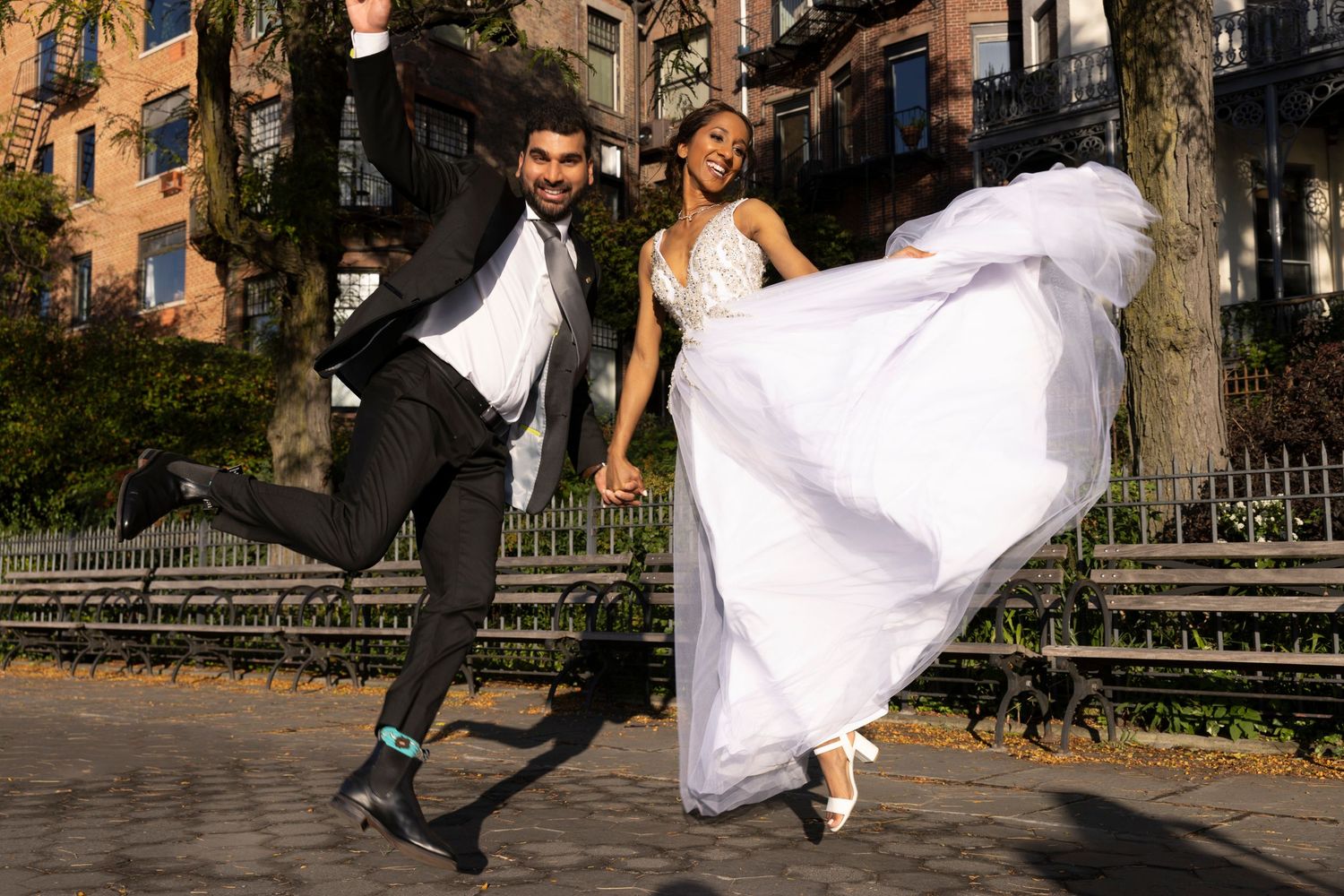 Couple in wedding attire dancing and twirling together in an outdoor urban setting with fall foliage in the background.