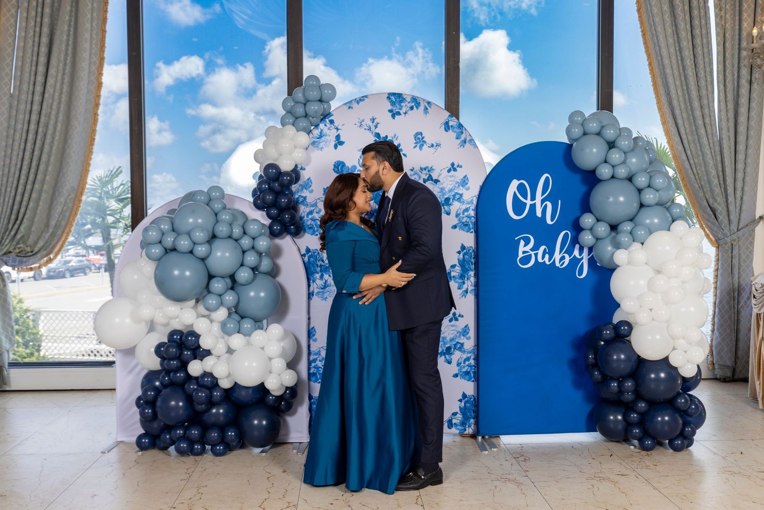 Elegant blue and white balloon arch decorations adorn a wedding venue with gray curtains and windows.