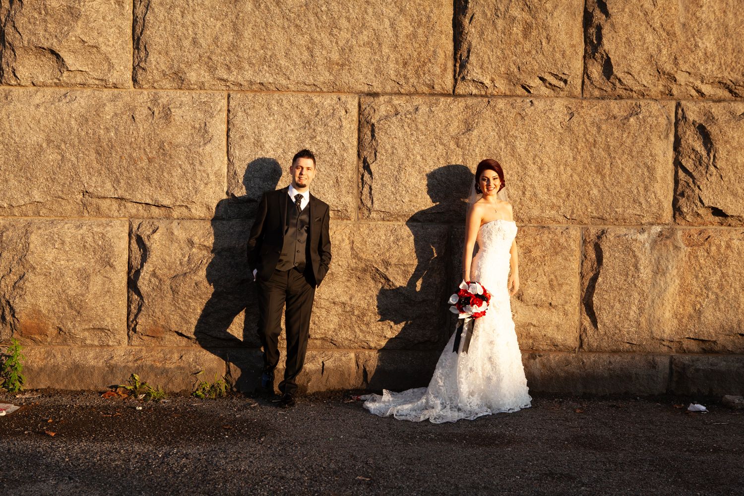 Wedding couple poses against rustic stone wall at sunset casting dramatic shadows.