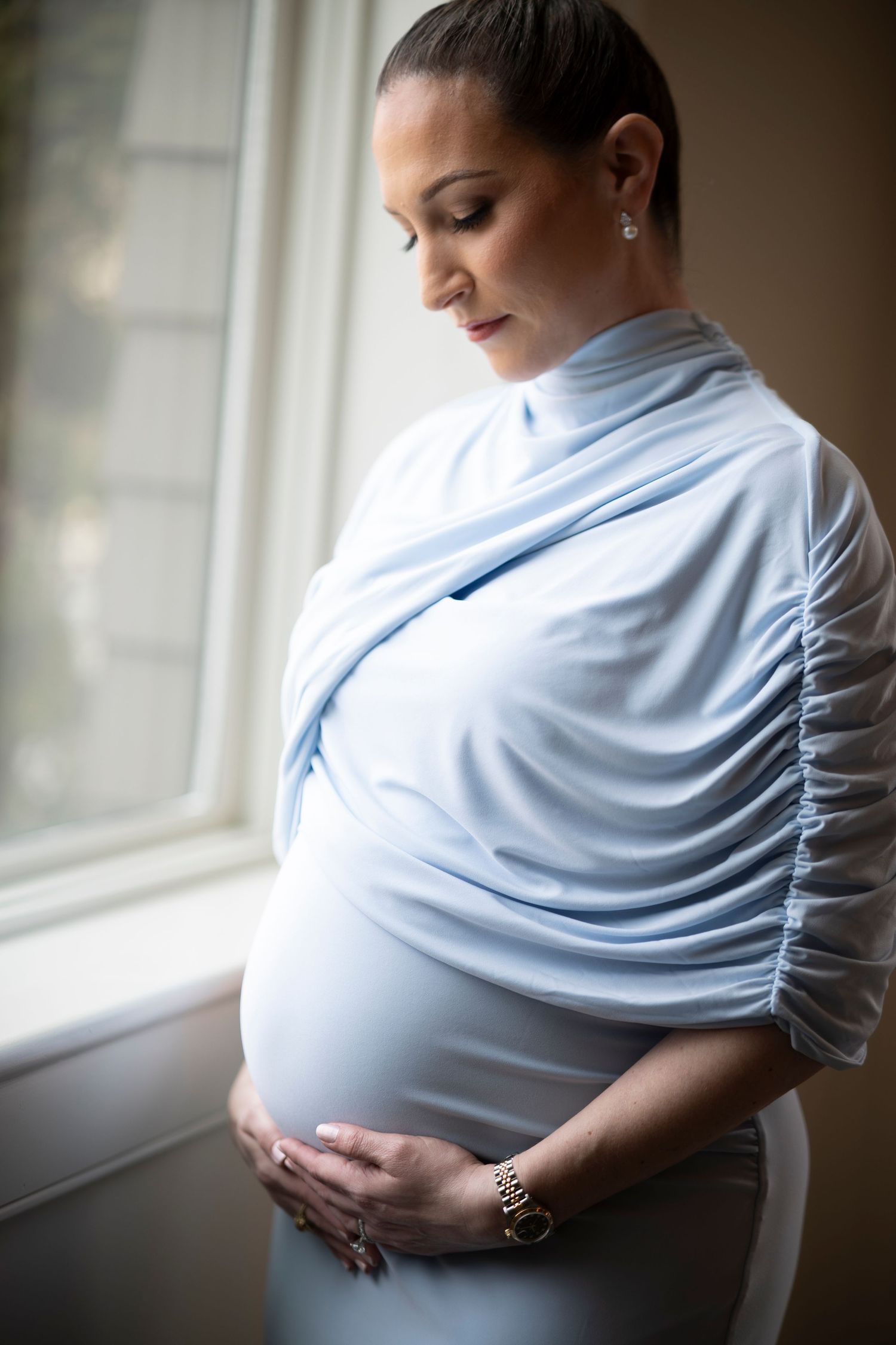 Expectant mother in soft blue wrap dress stands by window with hands gently cradling her pregnant belly.