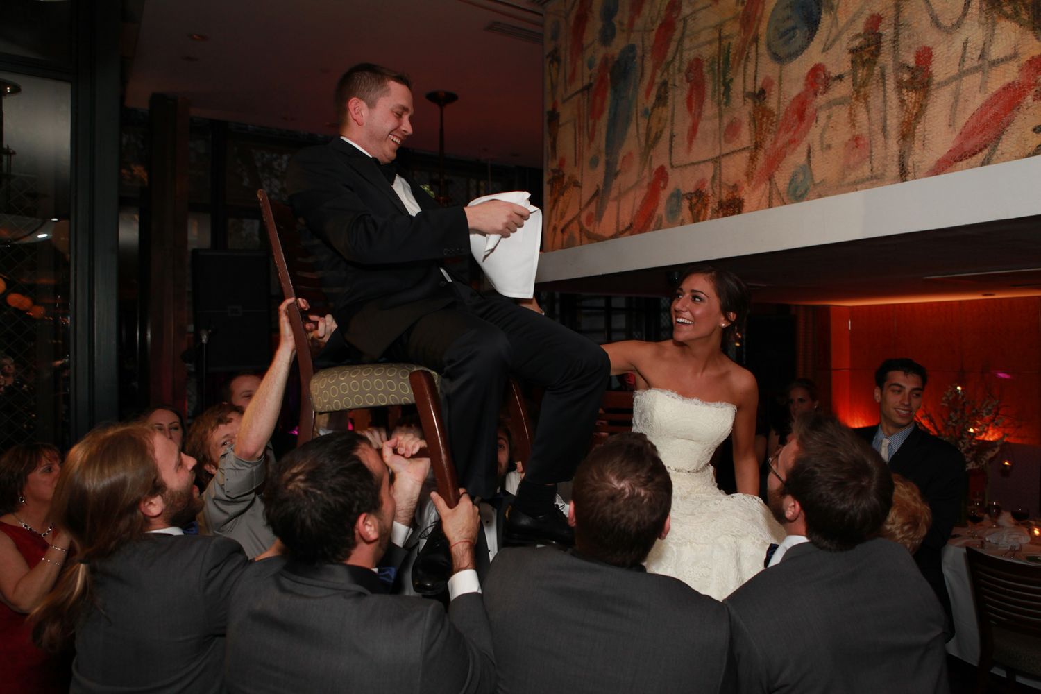 Wedding celebration with crowd lifting couple on chairs during traditional dance at reception.