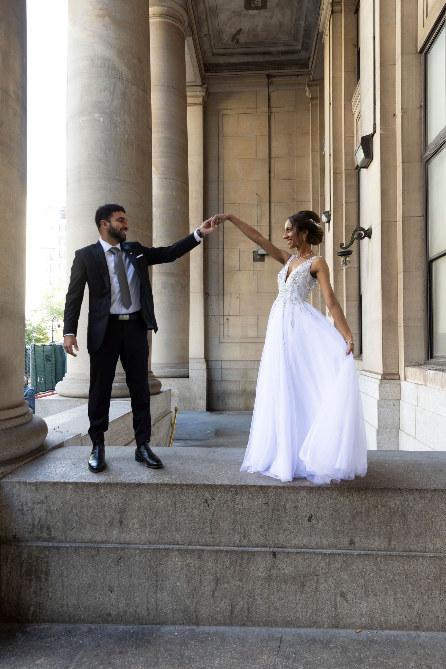 A couple in wedding attire dances playfully between elegant stone columns in a classic architectural setting.
