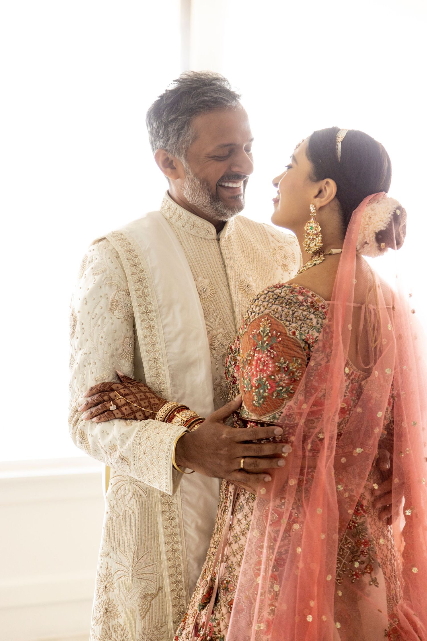 A couple in traditional Indian wedding attire share an intimate moment, wearing ornate cream and coral outfits with detailed embroidery.