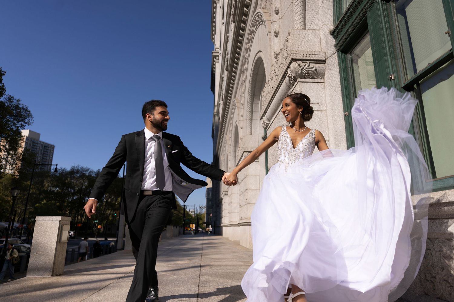 A wedding couple dances and twirls together in front of a grand stone church building on a sunny afternoon.