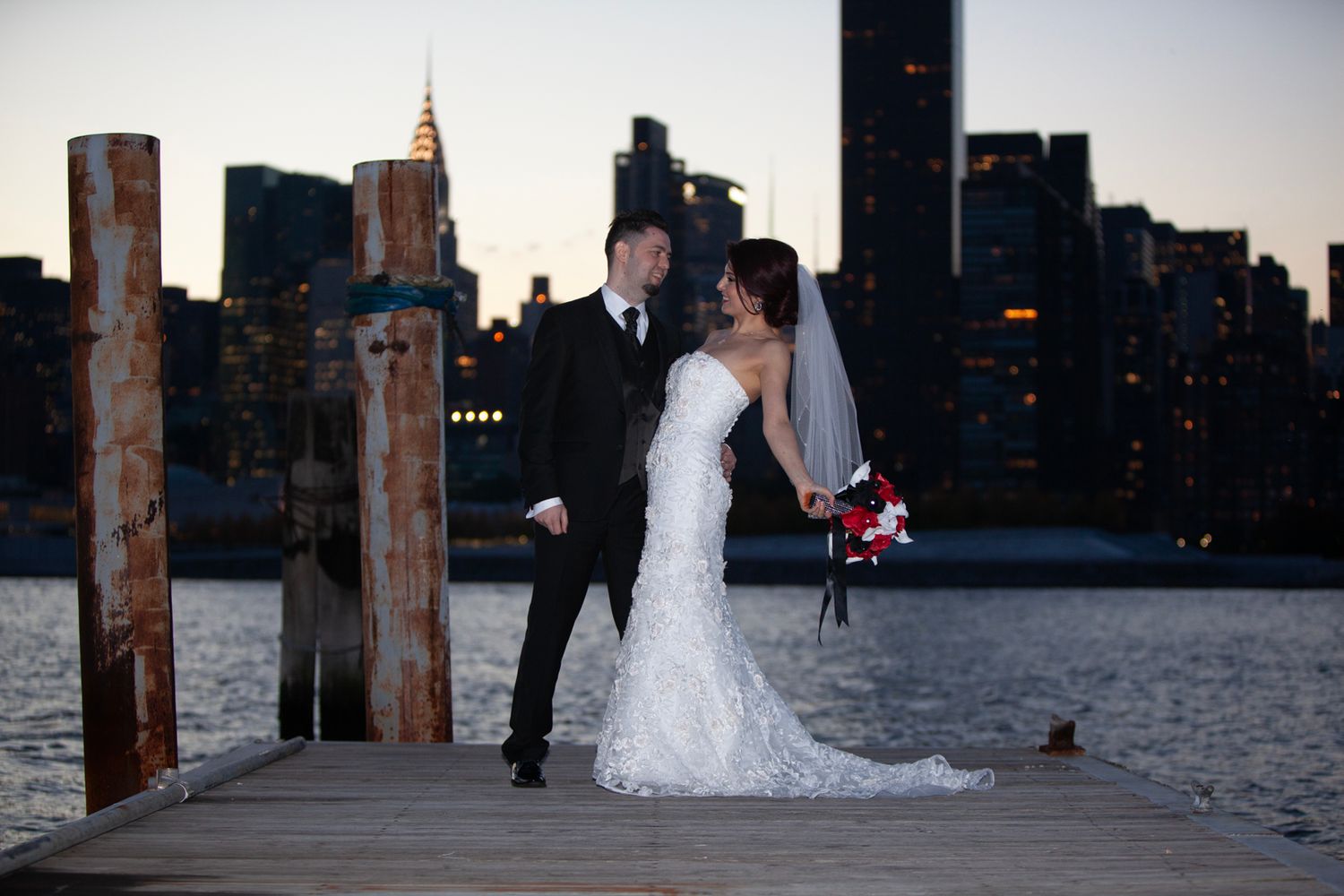 Couple in wedding attire sharing romantic moment on pier with Manhattan skyline at dusk.