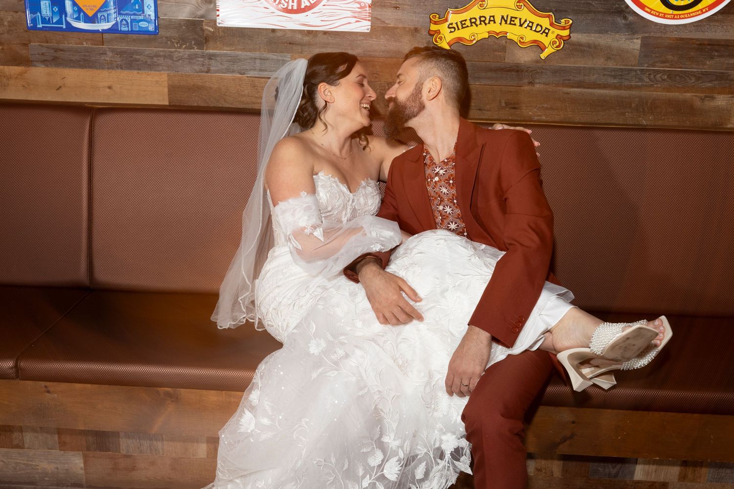 Bride and groom share a romantic moment seated together, bride in white lace gown with veil, groom in rust suit.