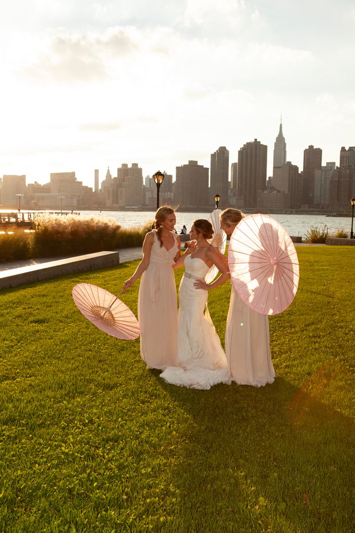 Three women in white dresses holding parasols with NYC skyline at sunset.