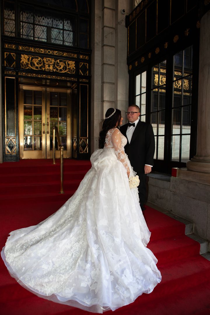 A bride in a flowing white gown poses dramatically on a red carpet.
