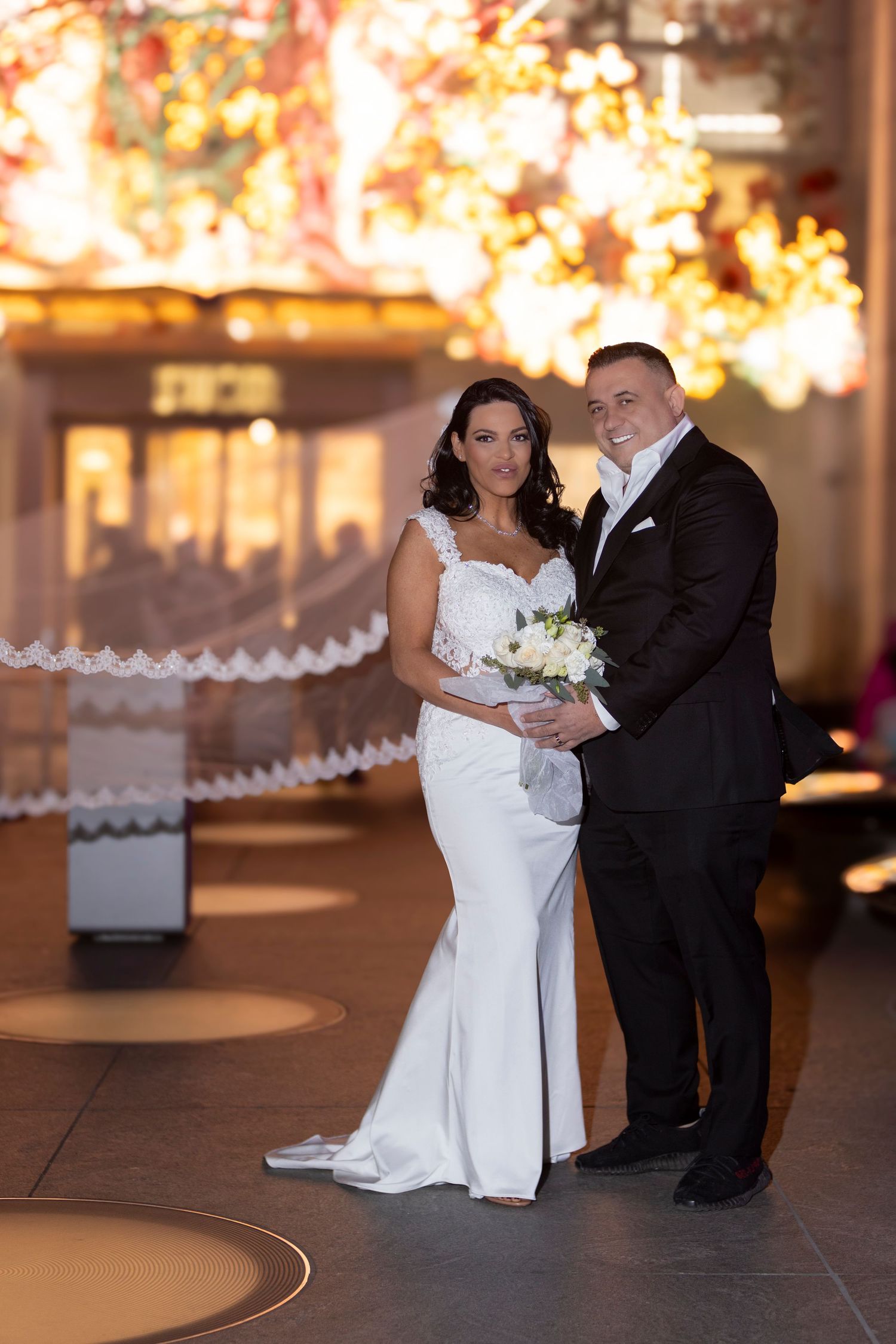 A wedding couple poses in formal attire against a backdrop of glowing holiday lights.