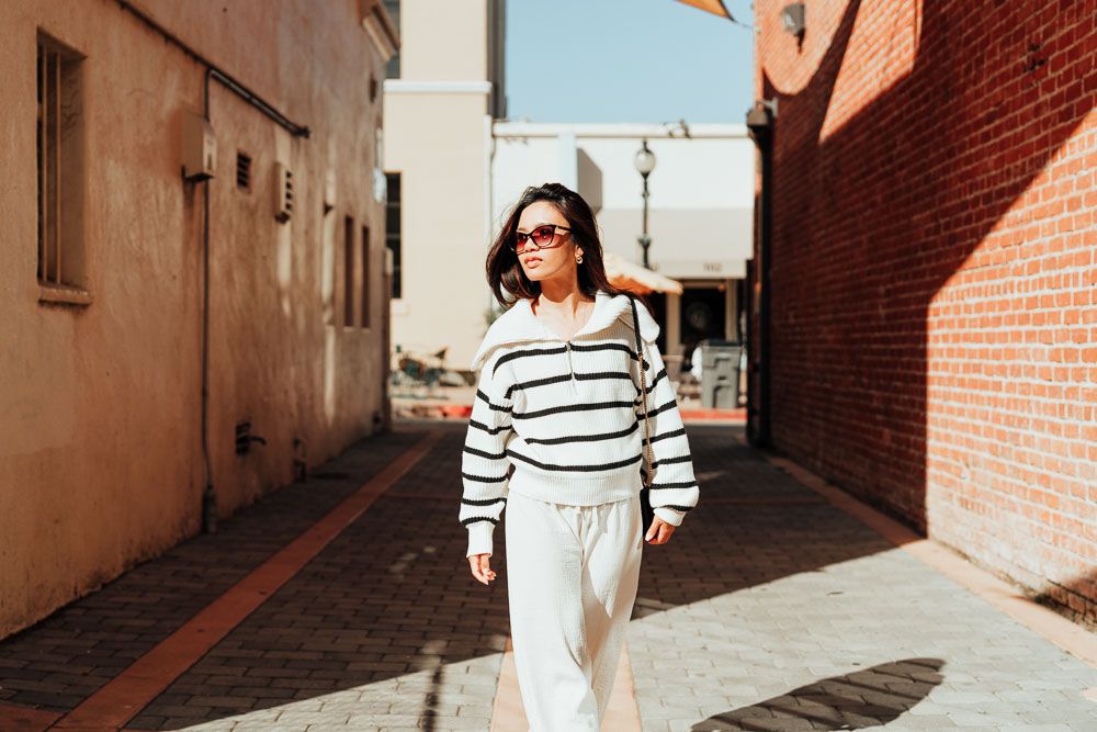 Street fashion photo of person in striped sweater and white pants walking in urban alley.