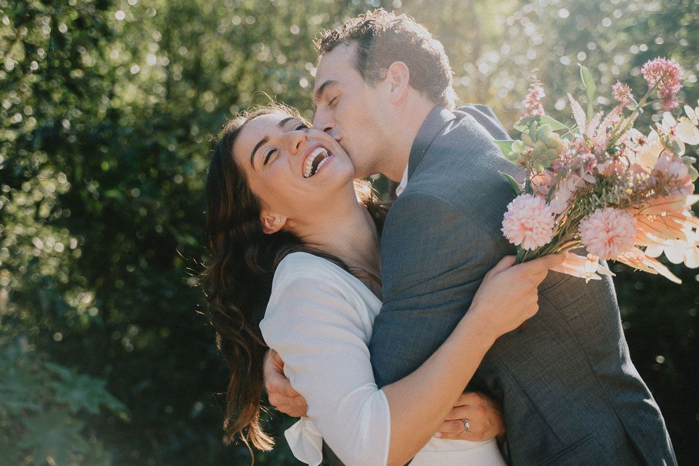Joyful couple laughing while holding a pink floral bouquet in a sunlit garden setting.