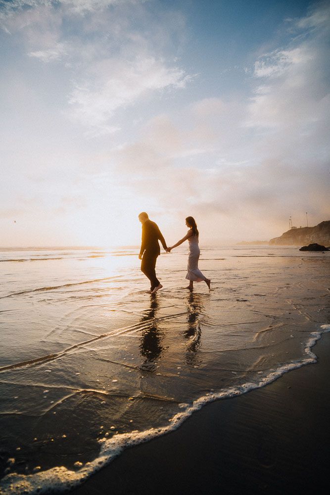 Two figures walking along the shoreline at golden hour with ocean waves lapping at their feet.