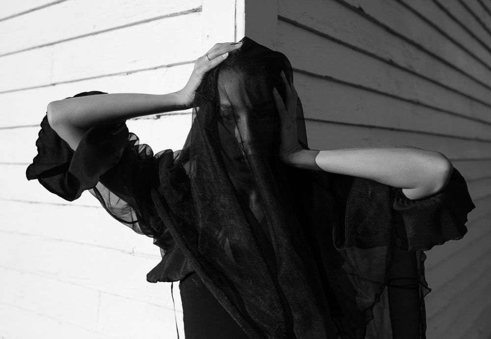 Dramatic black and white photo of figure with sheer black fabric against white wall.