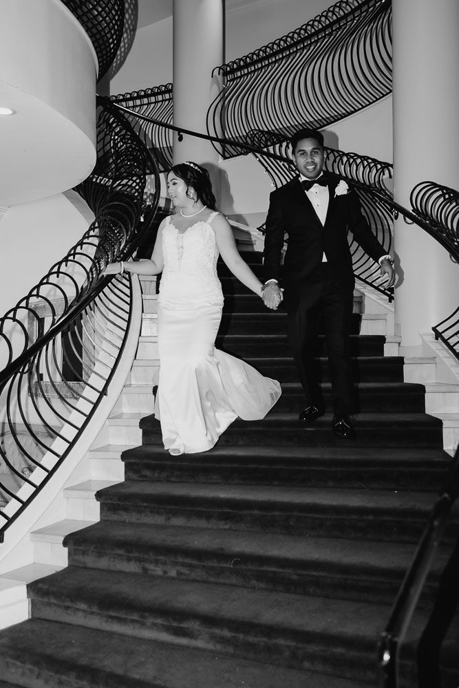 Elegant black and white photo of a couple descending a grand staircase with ornate railings.