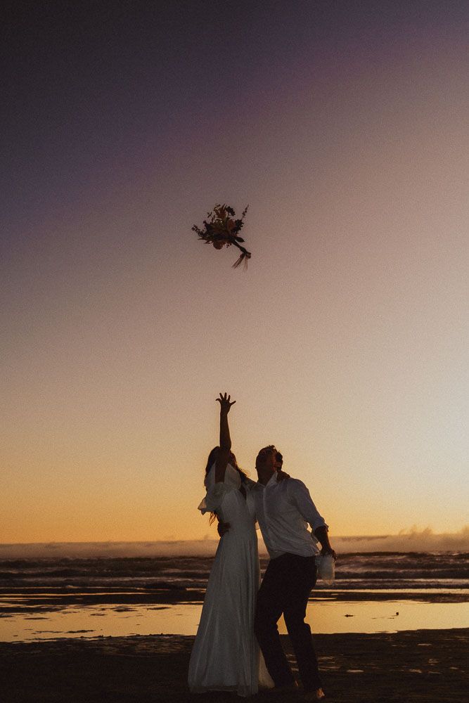 Silhouettes toss bouquet against sunset sky at beach.
