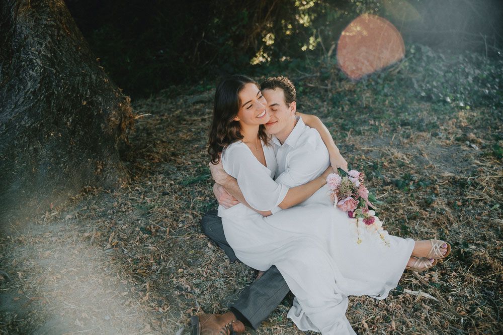 Intimate moment between couple in wedding attire sitting on rocky terrain.