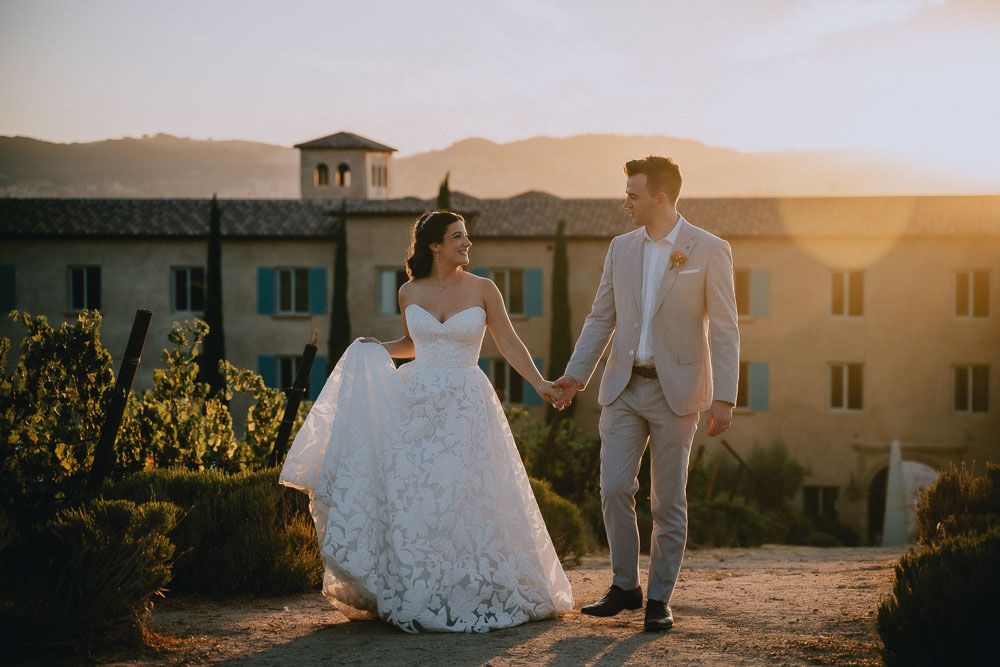 Newlyweds walking hand in hand at sunset with Mediterranean-style architecture in background.