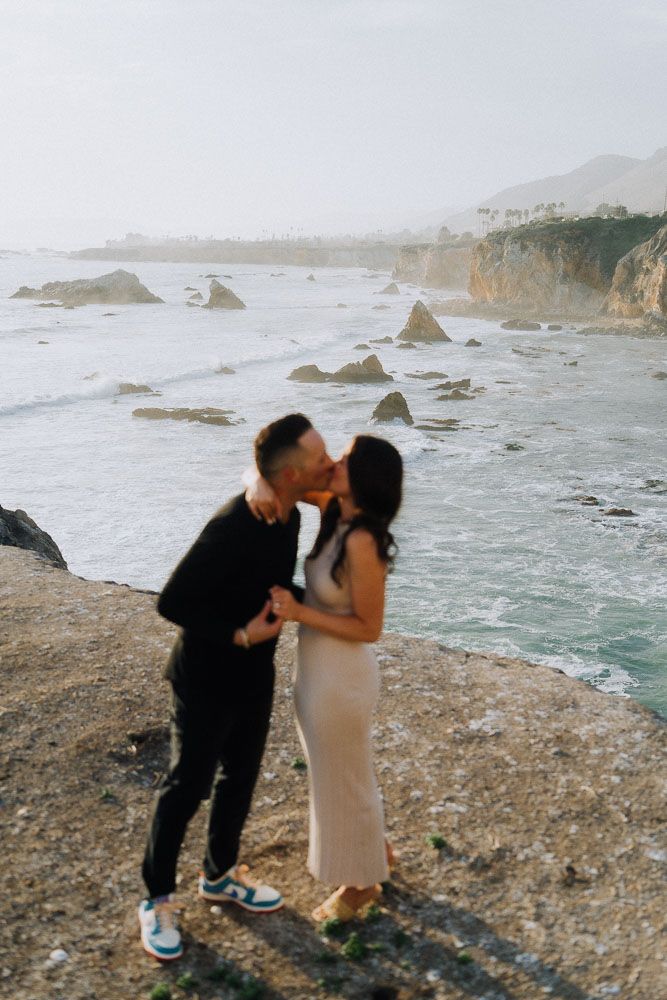 A tender moment shared on a rocky coastal overlook with crashing waves in the background.