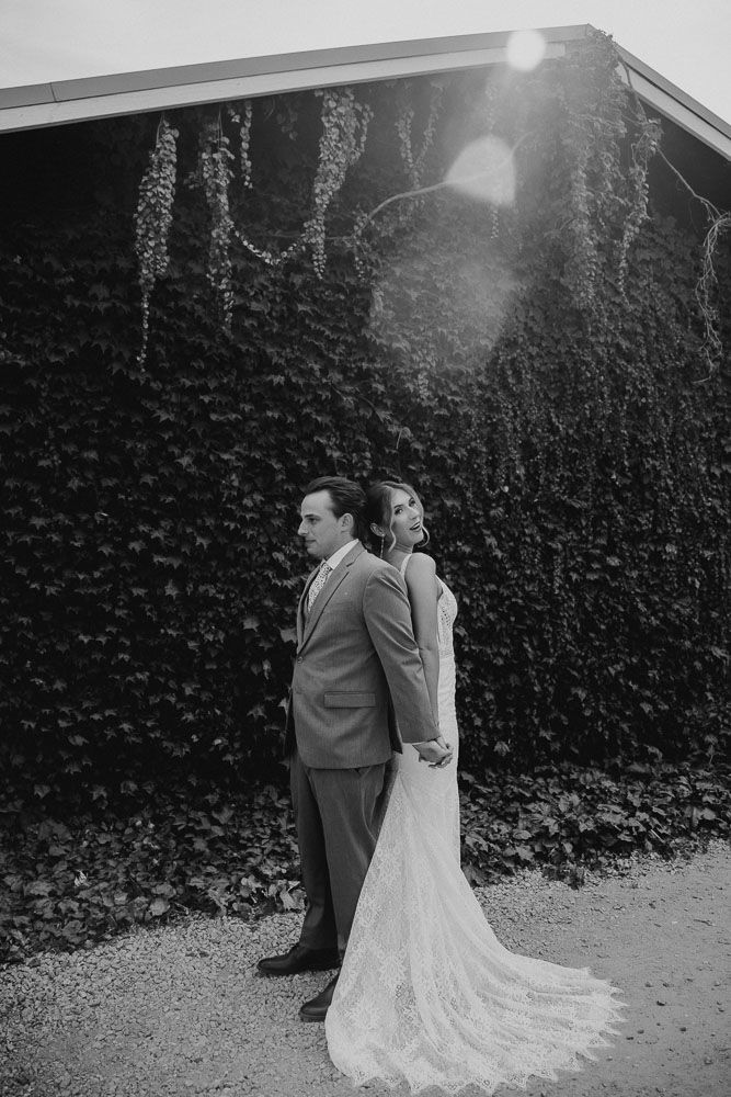A couple in wedding attire stand back-to-back against an ivy-covered wall in black and white.