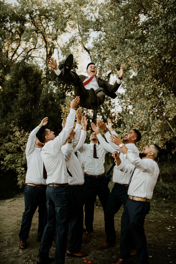 Groomsmen toss groom in the air during fun wedding photo in wooded setting.