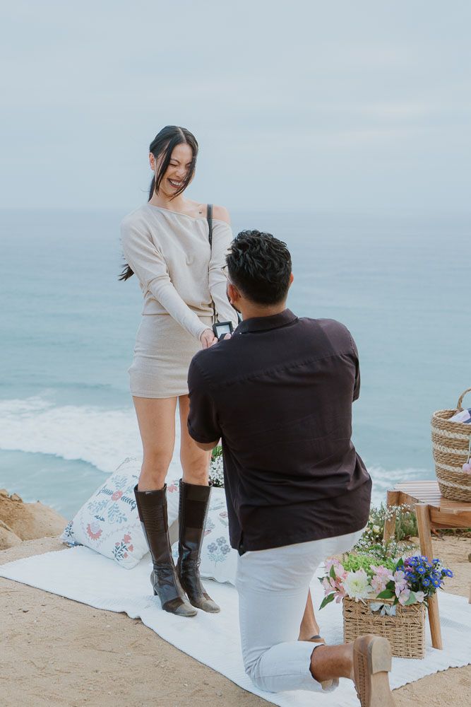Romantic proposal setup on coastal cliff with flowers and picnic blanket.